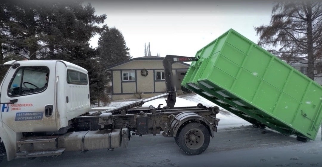 Hauling Heroes roll-off truck lowering a green bin at a residential property in winter