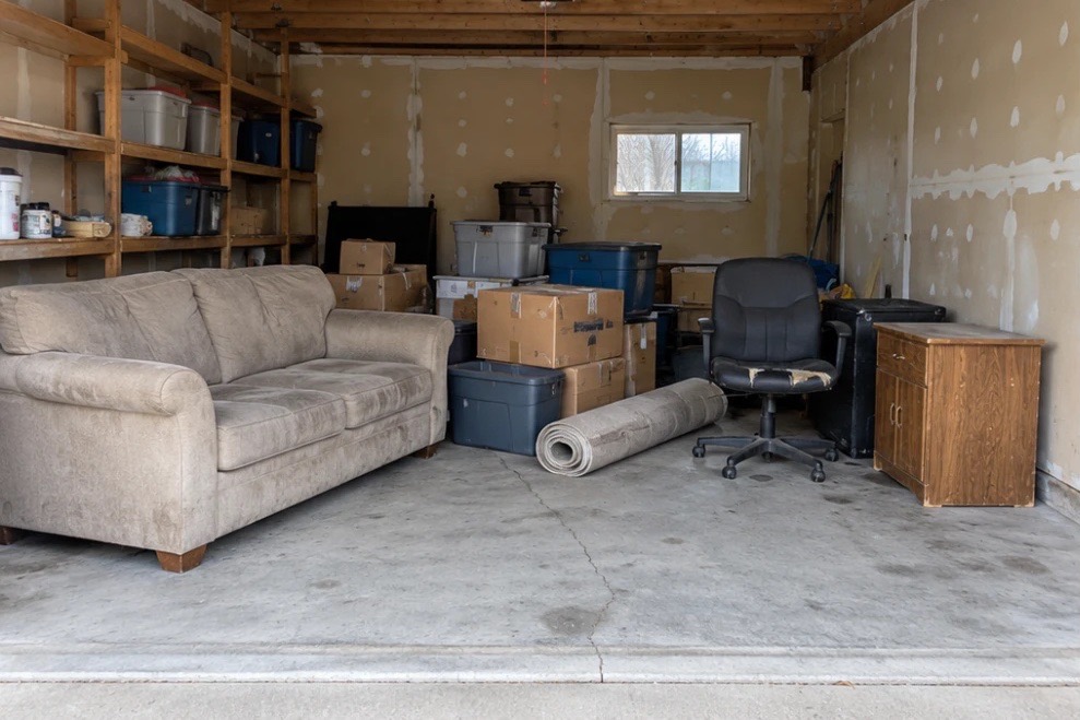 Single-car garage crowded with a couch, storage bins, boxes, an office chair, a rolled rug and a wooden cabinet