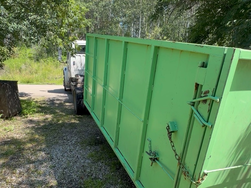 Green Hauling Heroes roll-off bin placed on a gravel lot beside trees
