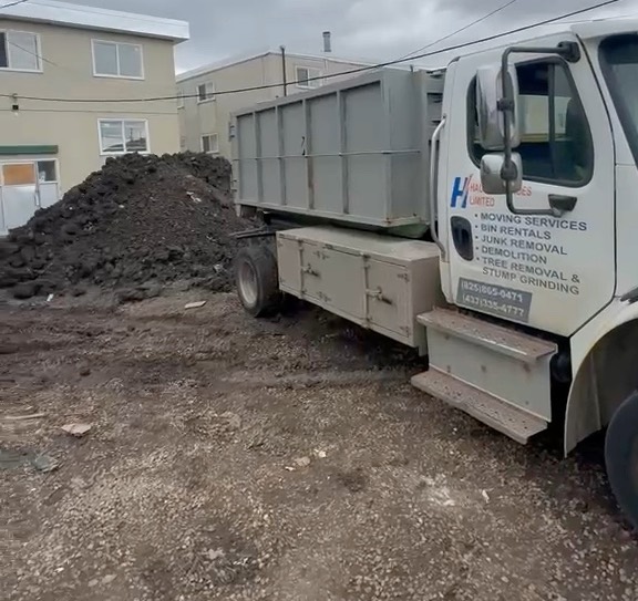 Hauling Heroes truck carrying a grey roll-off bin at a job site beside a pile of soil