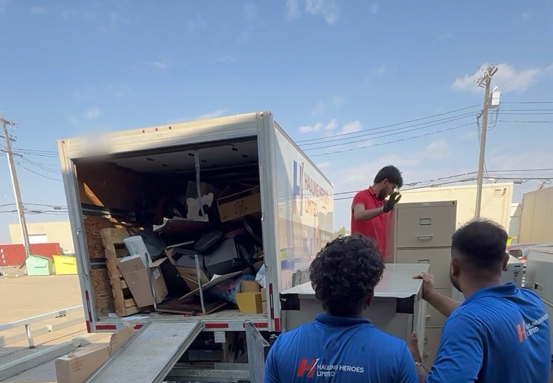 Two Hauling Heroes crew members in branded blue shirts loading office furniture and filing cabinets into a truck
