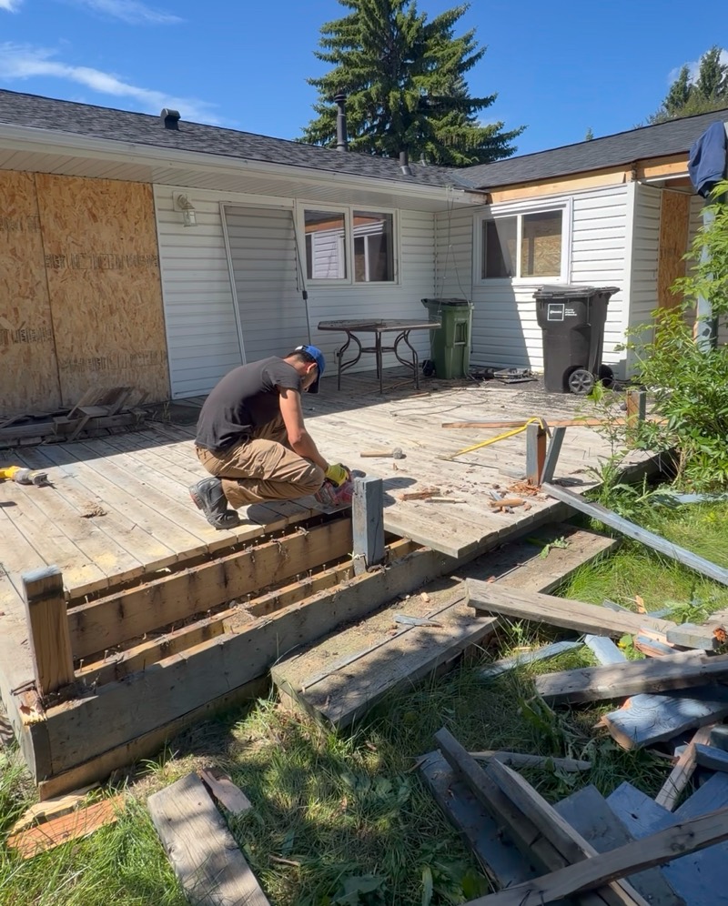 Crew member dismantling a backyard wooden deck, with removed boards stacked on the lawn