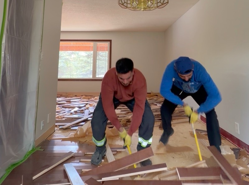 Two crew members prying up hardwood flooring in a living room, with doorways covered in plastic sheeting