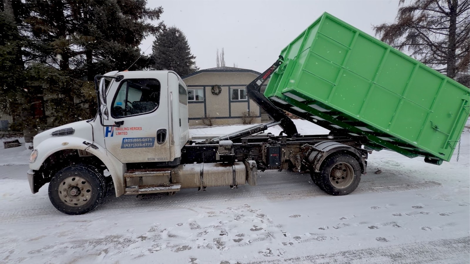 Hauling Heroes truck lifting a green roll-off bin on a snowy residential street