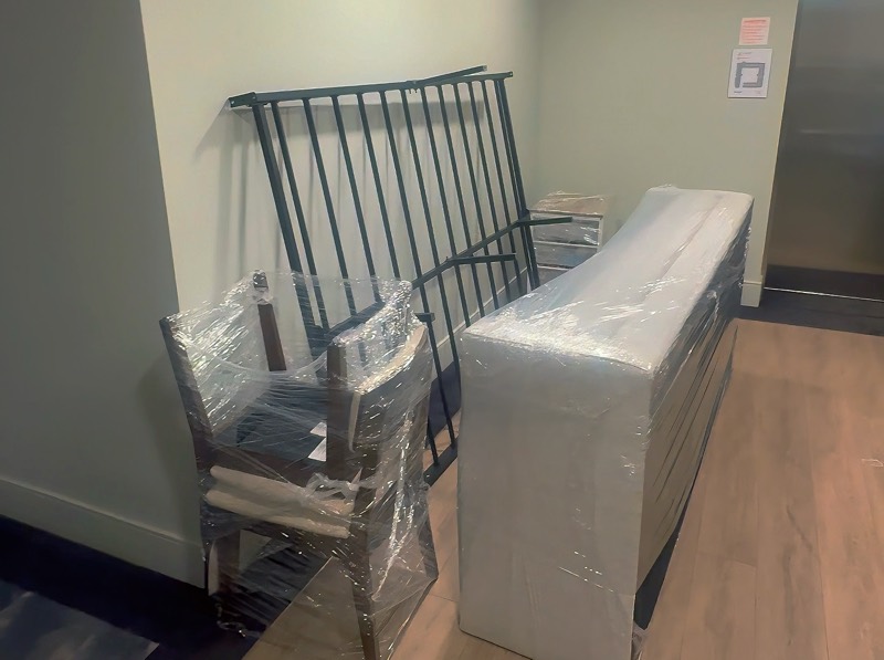 Shrink-wrapped chairs and a padded, plastic-wrapped sofa staged in an apartment hallway for a move